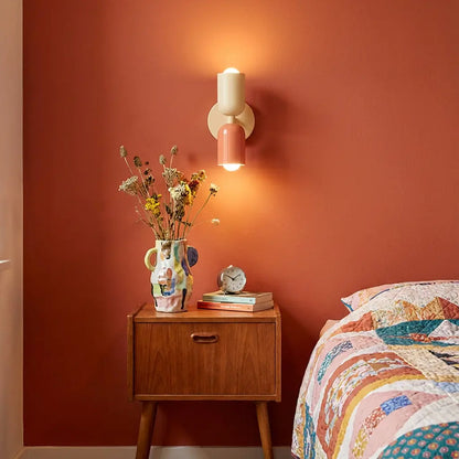 Leuchtende Wandlampe in Creme und Lachsrosa an terrakottafarbener Wand über einem Holz-Nachttisch mit bunter Vase und Trockenblumen. 1