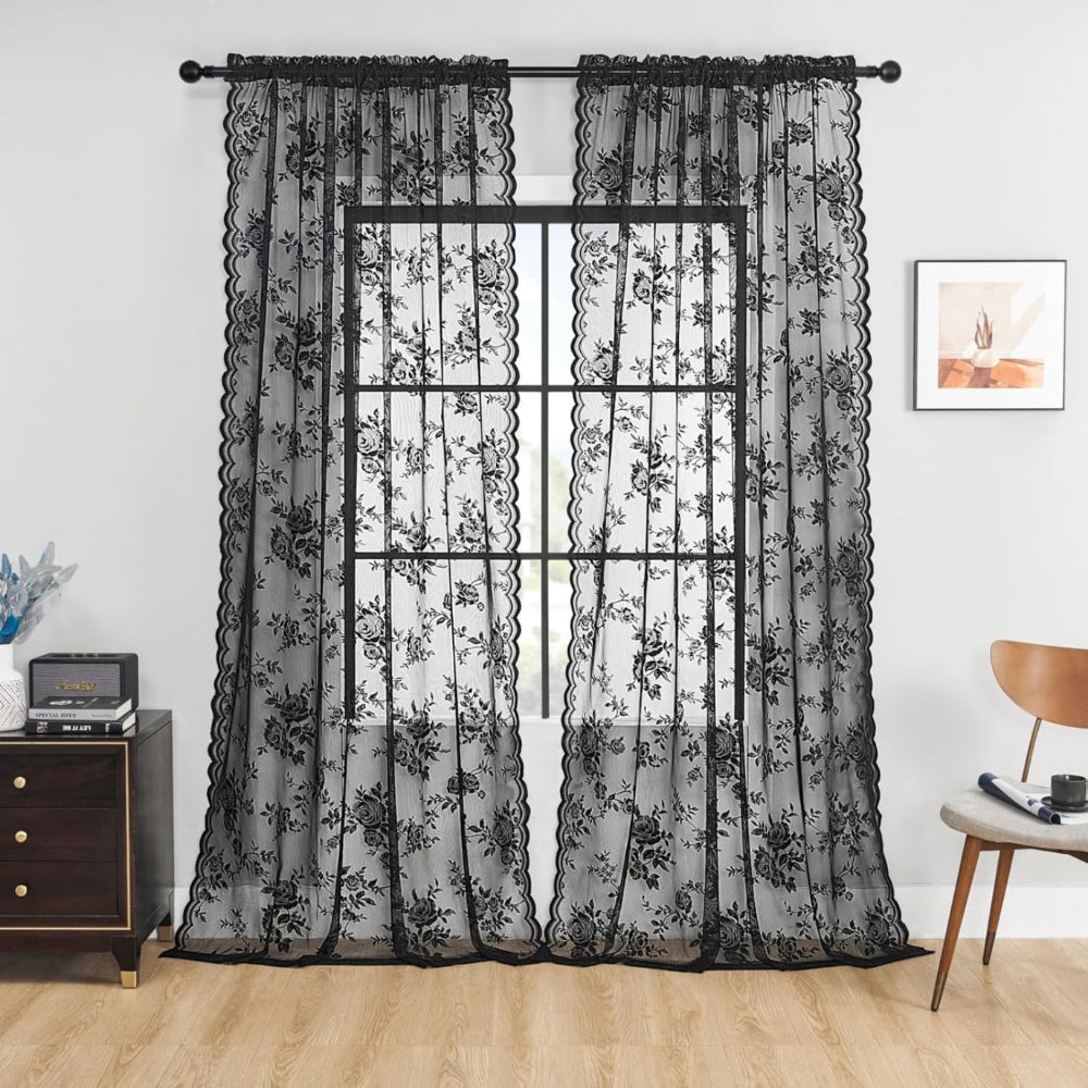 Zwei schwarze transparente Spitzenpaneele mit Rosenmuster und Wellenkante hängen an einer Stange vor einem Fenster in einem Wohnzimmer. 1