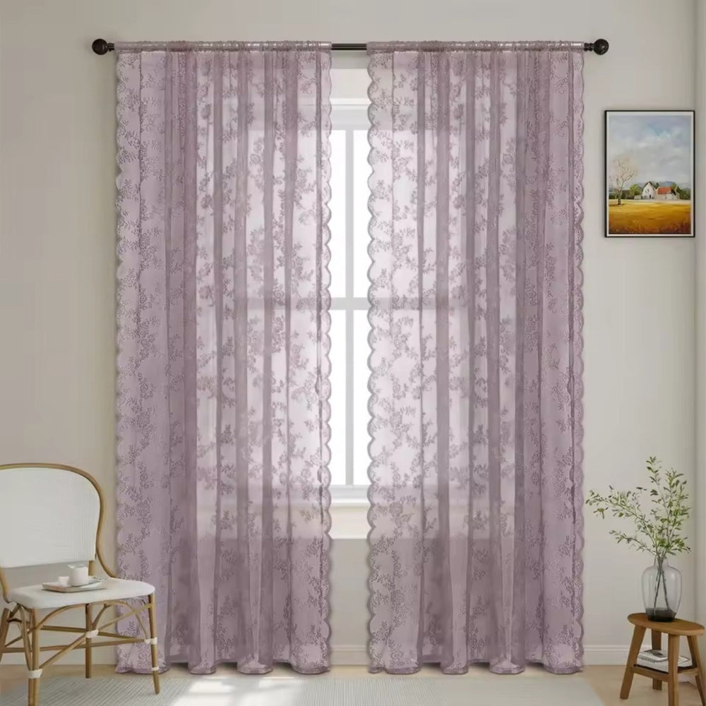 Zwei staubige malvenfarbene Spitzenpaneele mit Blumenmuster und Wellenkante hängen an einer Stange vor einem Fenster in einem hellen Zimmer. 11