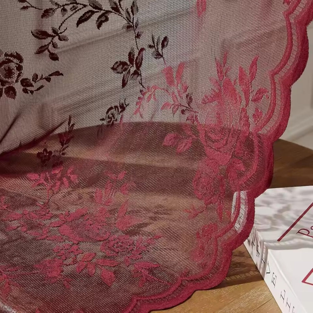 Nahaufnahme der weinroten Spitze mit aufgesticktem Rosenmuster und gewellter Kante, die über einer Holzfläche neben einem Buch liegt. 3