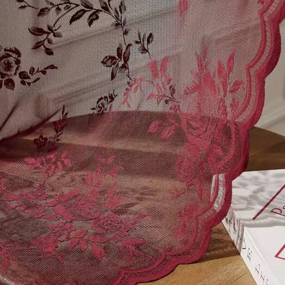 Nahaufnahme der weinroten Spitze mit aufgesticktem Rosenmuster und gewellter Kante, die über einer Holzfläche neben einem Buch liegt. 3
