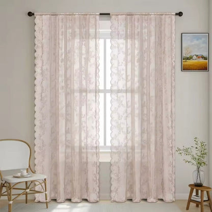 Zwei zartrosa transparente Spitzenpaneele mit feinem Blütenmuster und Wellenkante vor einem Fenster, daneben ein Rattanstuhl und ein Holzhocker mit Pflanze. 9