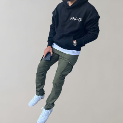 Mann in schwarzem Hoodie, olivgrüner Cargohose, weißen Sneakers, hält Smartphone.