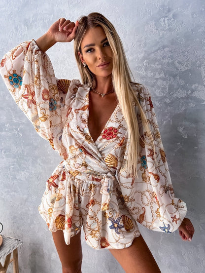 Frau in weißem, gemustertem Sommerkleid mit Muschel- und Kettenmotiven vor grauer Wand.