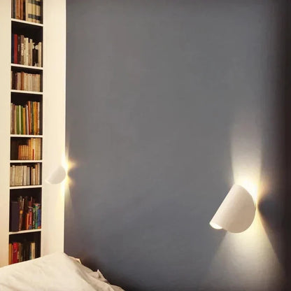 Zwei weiße Wandleuchten an dunkler Wand neben einem Bücherregal über dem Bett.