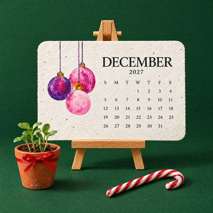 Dezemberblatt mit rosa und violetten Christbaumkugeln auf Staffelei, davor ein Zuckerstange und ein Töpfchen mit Keimlingen 3