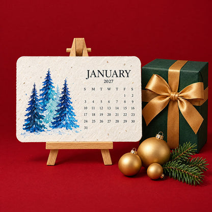 Januarblatt mit blauen Aquarelltannen auf Holzstaffelei, daneben ein grünes Geschenk mit Goldschleife und goldene 2