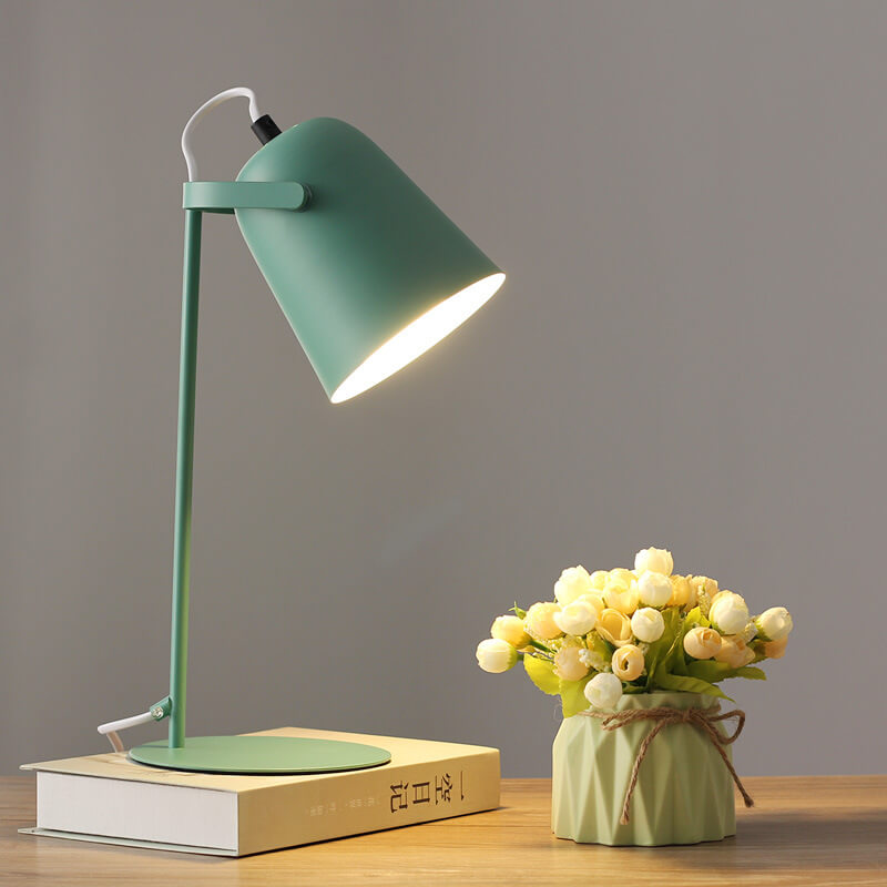Grüne Tischlampe leuchtet auf einem Buch, daneben ein Väschen mit cremefarbenen Röschen vor grauer Wand. 9