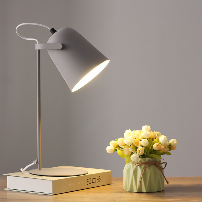 Graue Tischlampe mit eimerförmigem Schirm brennt auf einem Buch, daneben ein kleiner Blumenstrauß im grünen Väschen. 10