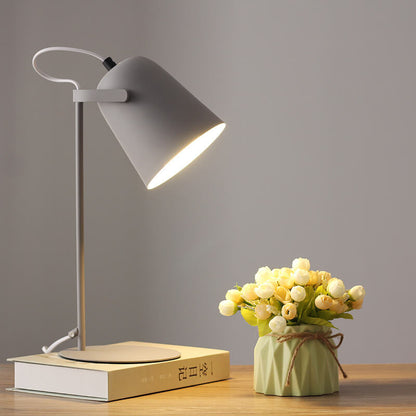 Graue Tischlampe mit eimerförmigem Schirm brennt auf einem Buch, daneben ein kleiner Blumenstrauß im grünen Väschen. 10