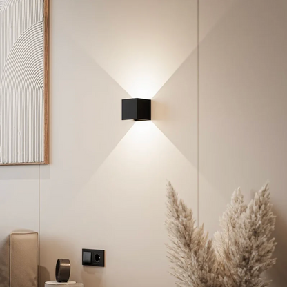 Schwarze würfelförmige Wandlampe an heller Wand, die Licht nach oben und unten wirft, daneben Pampasgras und Steckdose. 1