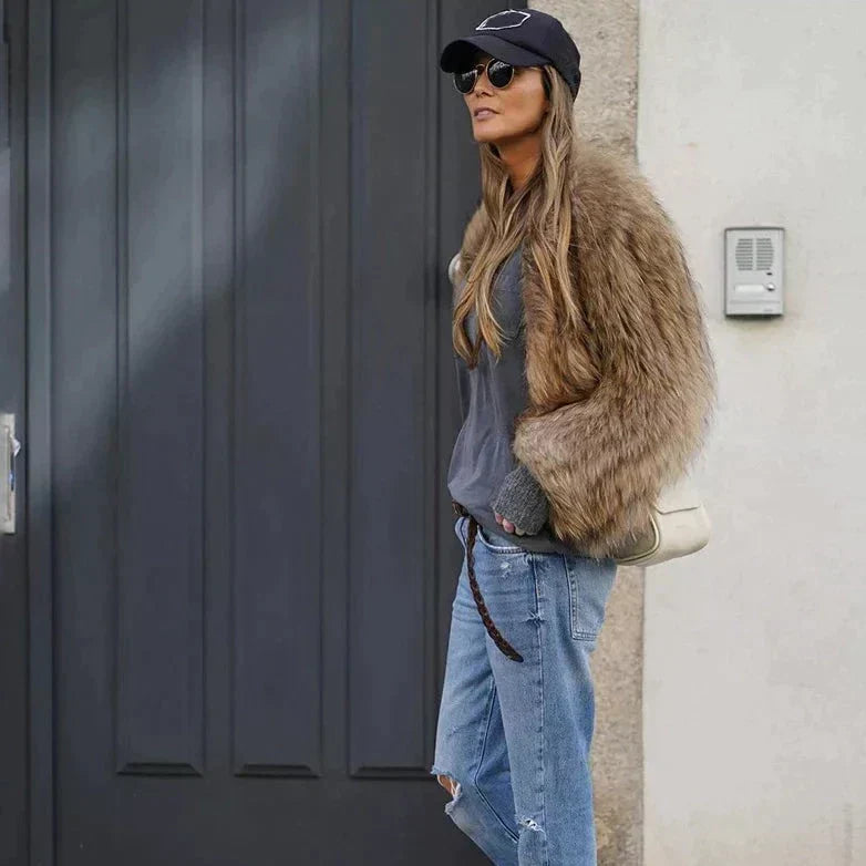 Frau in brauner Kunstpelzjacke, blauer Jeans und schwarzer Kappe vor grauer Tür.