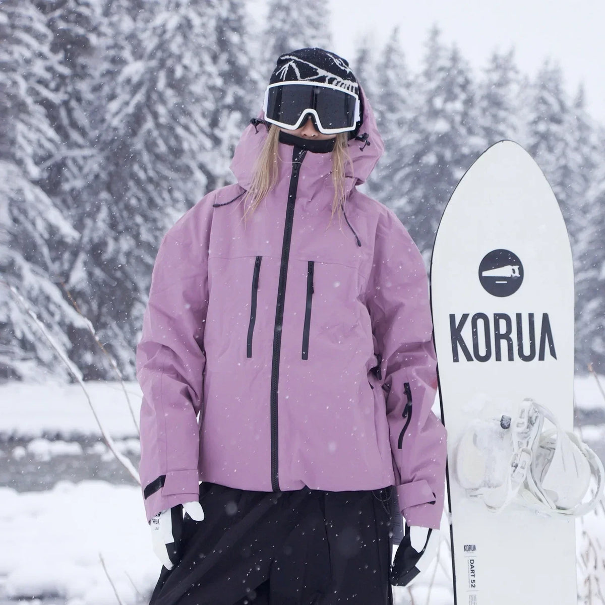 Rosa Skijacke mit schwarzen Reißverschlüssen frontal getragen, daneben steht ein weißes Snowboard mit Bindung im Schnee. 3