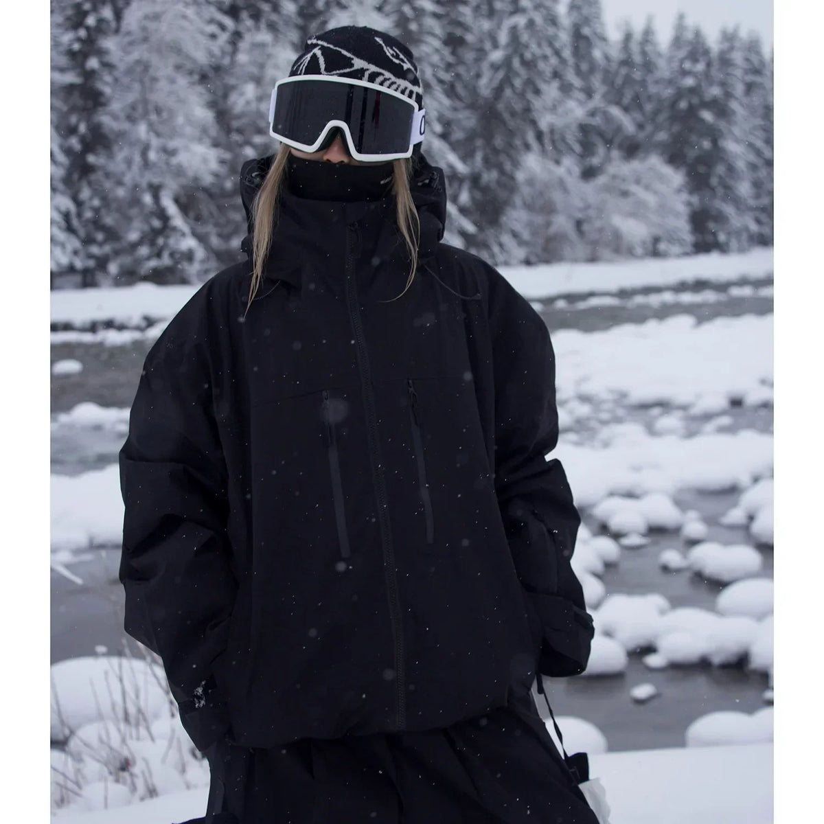 Schwarze Skijacke mit Kapuze und hohem Kragen an einer Person mit Skibrille, im Schneefall vor einem Bach. 2