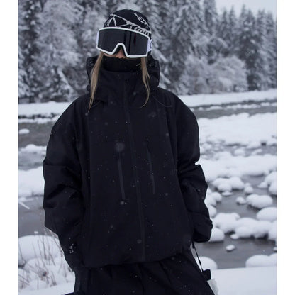 Schwarze Skijacke mit Kapuze und hohem Kragen an einer Person mit Skibrille, im Schneefall vor einem Bach. 2