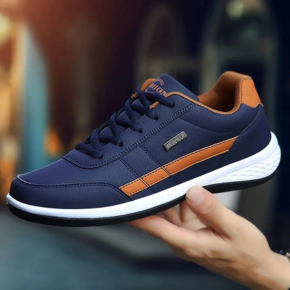 Dunkelblauer Ledersneaker mit orangebraunem Seitenpaneel und weißer Sohle, von einer Hand seitlich präsentiert.