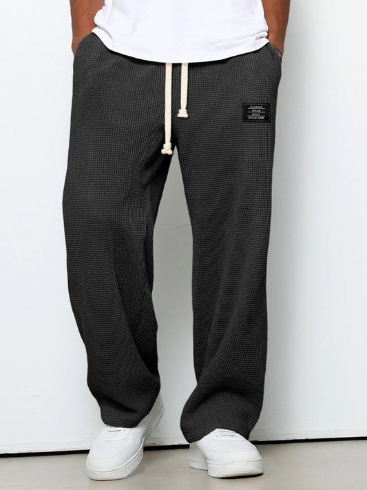 Frontansicht der Waffelstrick-Hose in Anthrazit, getragen von einem Model mit weißem T-Shirt und weißen Sneakern, Hände in den Taschen. 1