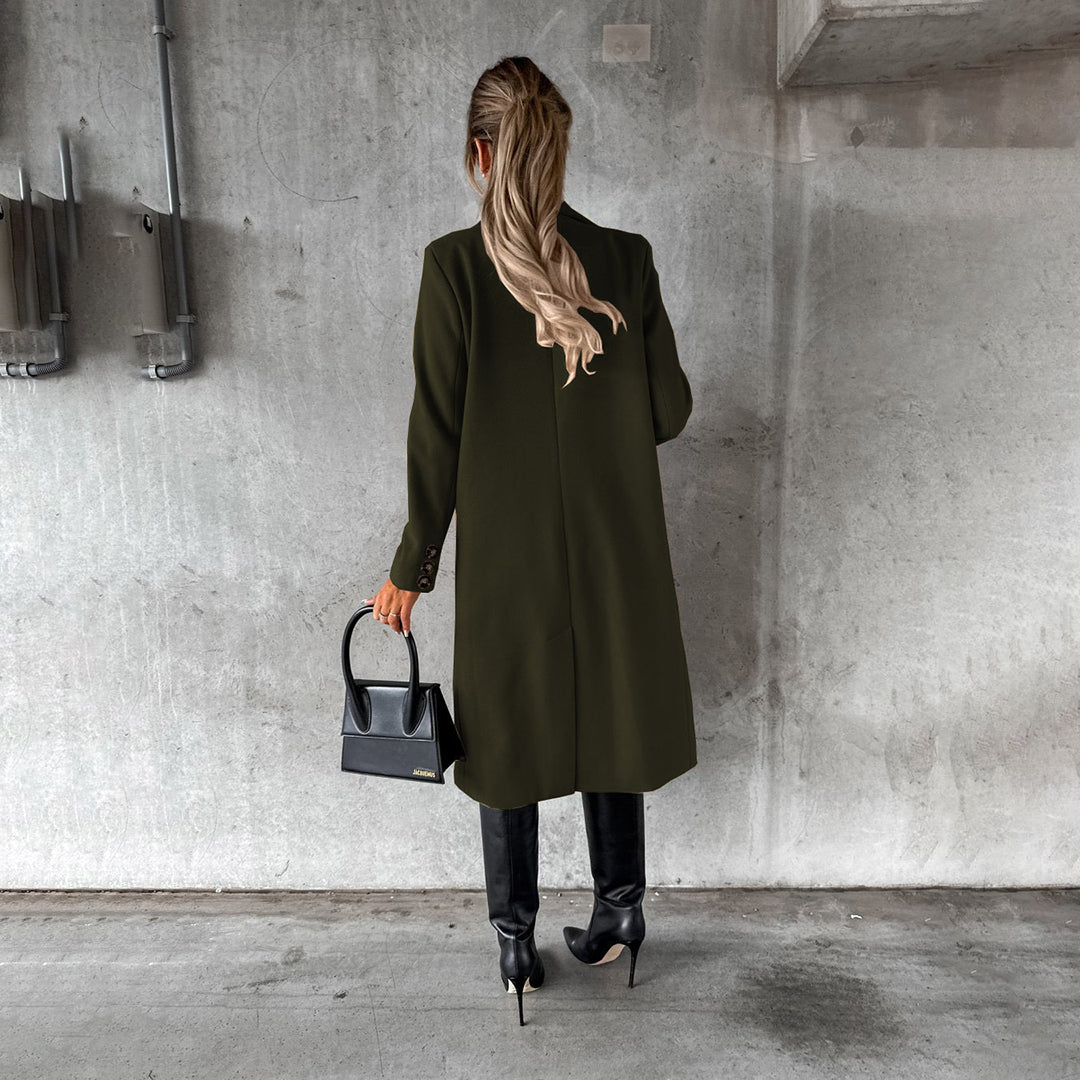 Frau in olivgrünem Wintermantel, schwarze Stiefel, schwarze Handtasche, Betonhintergrund.