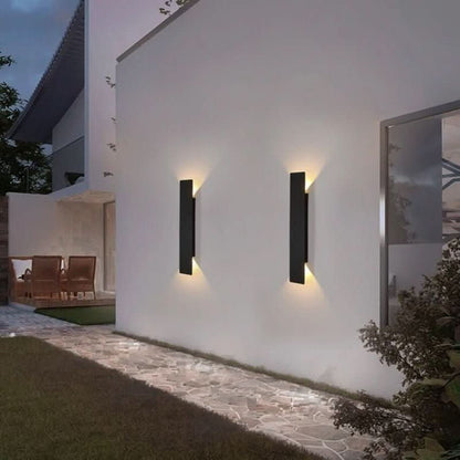 Zwei schwarze zylindrische LED-Wandleuchten an einer weißen Hauswand im Garten.