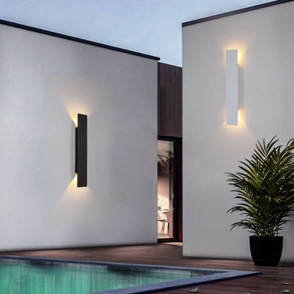 Eine schwarze und eine weiße zylindrische LED-Wandleuchte an hellen Außenwänden neben einem Pool.