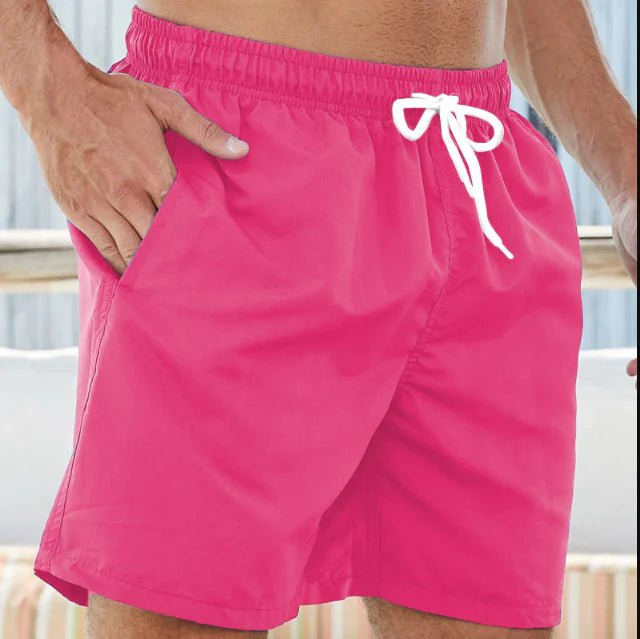 Pinke Herren-Badeshorts mit weißer Kordel, getragen, mit Strandkulisse im Hintergrund.