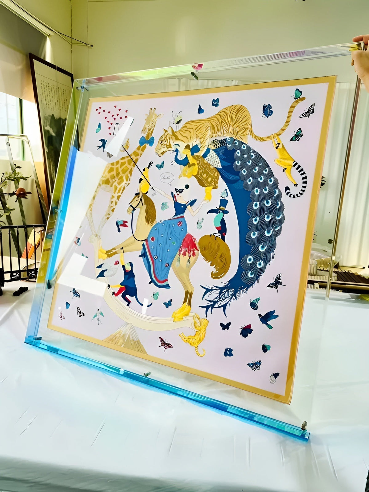 Großer Acrylrahmen mit blauer Kante, von einer Hand gehalten, darin ein Seidentuch mit Tieren und Schmetterlingen