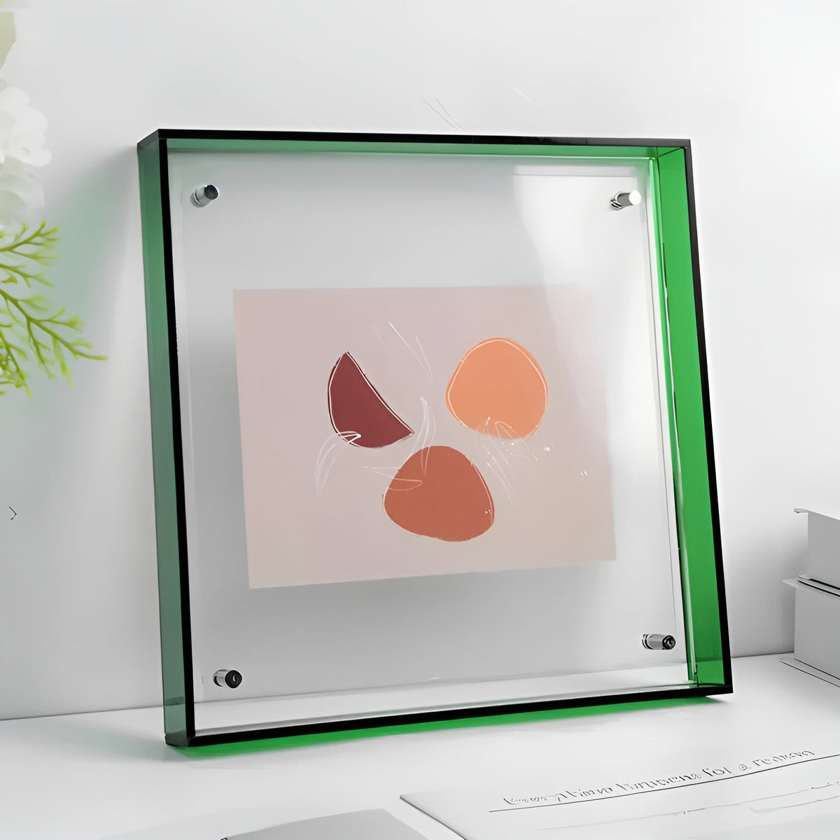 Quadratischer Acrylrahmen mit grünem Rand und sichtbaren Haltebolzen, steht auf einem weißen Tisch und zeigt eine abstrakte Grafik in Orange- und Rottönen. 21