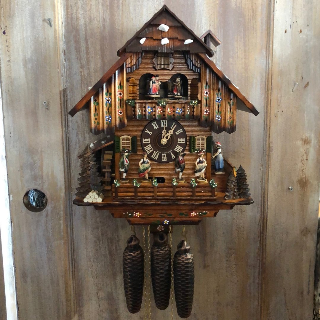 Aufgehängte Kuckucksuhr an einer verwitterten Holzwand, mit Blumenbemalung am Dach, Tanzfiguren, Wasserrad und drei dunklen Zapfengewichten. 12