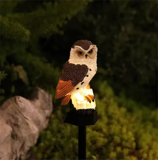 Braun-weiße Eule mit orangefarbenen Flügelspitzen, abends leuchtend auf Spieß, im Hintergrund Gebüsch und ein Stein. 7