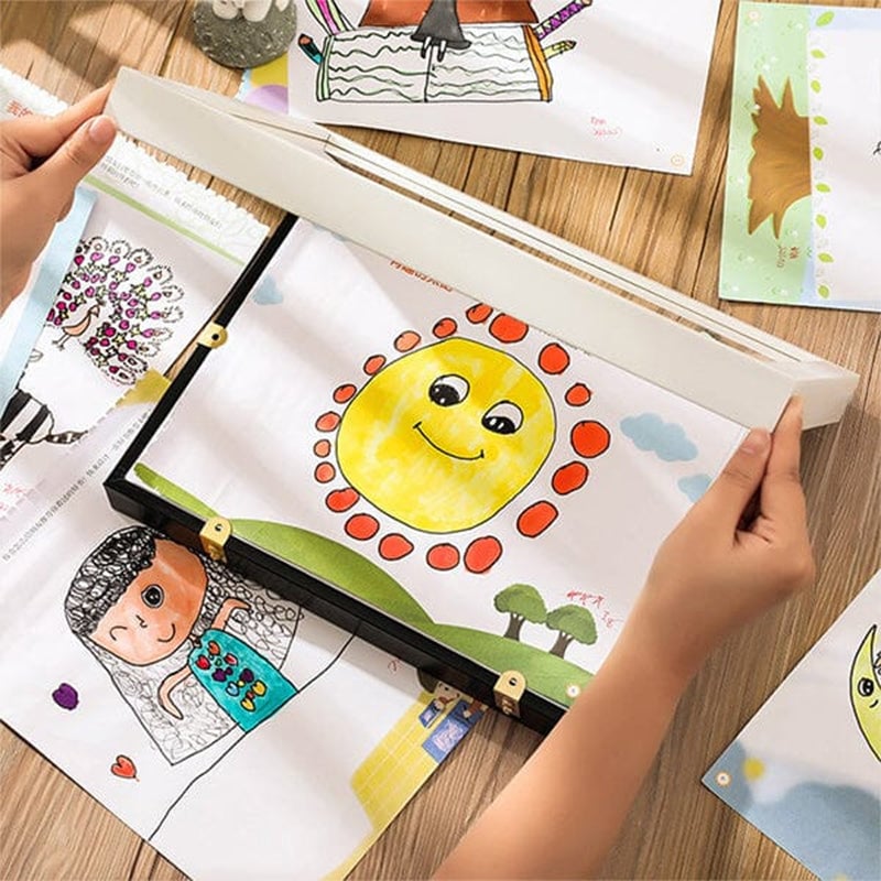 Hände legen eine Kinderzeichnung mit lächelnder Sonne in ein flaches Rahmenfach auf einem Holztisch.