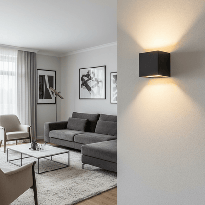 Schwarze Würfel-Wandlampe in Nahaufnahme an einer hellen Wand, im Hintergrund ein Wohnzimmer mit grauem Sofa und gerahmten Bildern. 2
