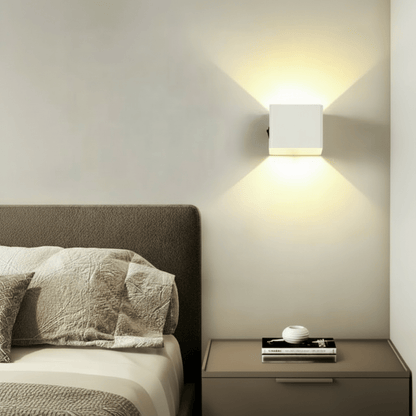 Weiße Würfel-Wandlampe über einem Nachttisch neben dem Bett, das warme Licht fällt nach oben und unten an die Wand. 3