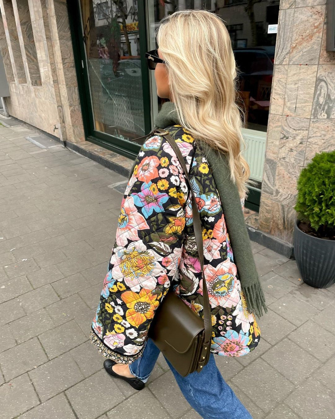 Frau mit floralem Steppmantel, grüner Tasche und Schal auf Gehweg.