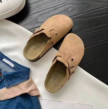 Beige Wildleder-Schlupfclogs auf schwarzem Holzboden, modisch und bequem.