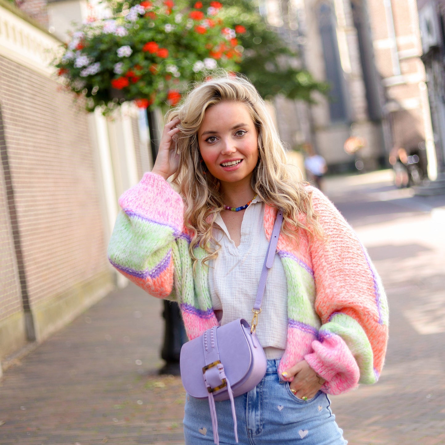 Frau in bunter Strickjacke, lila Tasche, Jeans mit Herzmuster, lächelnd auf Straße.