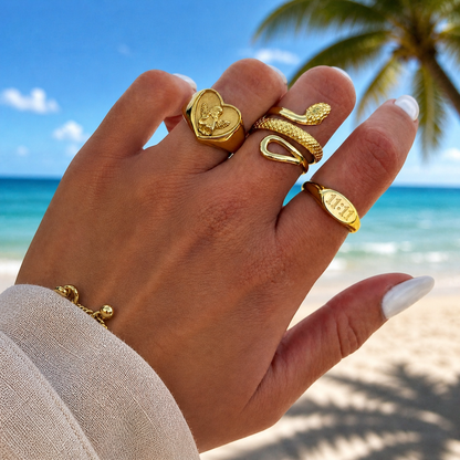 Hand am Strand mit Engelherz-Ring am Zeigefinger, kombiniert mit Schlangenring und ovalem Siegelring, dazu goldenes Armband. 3