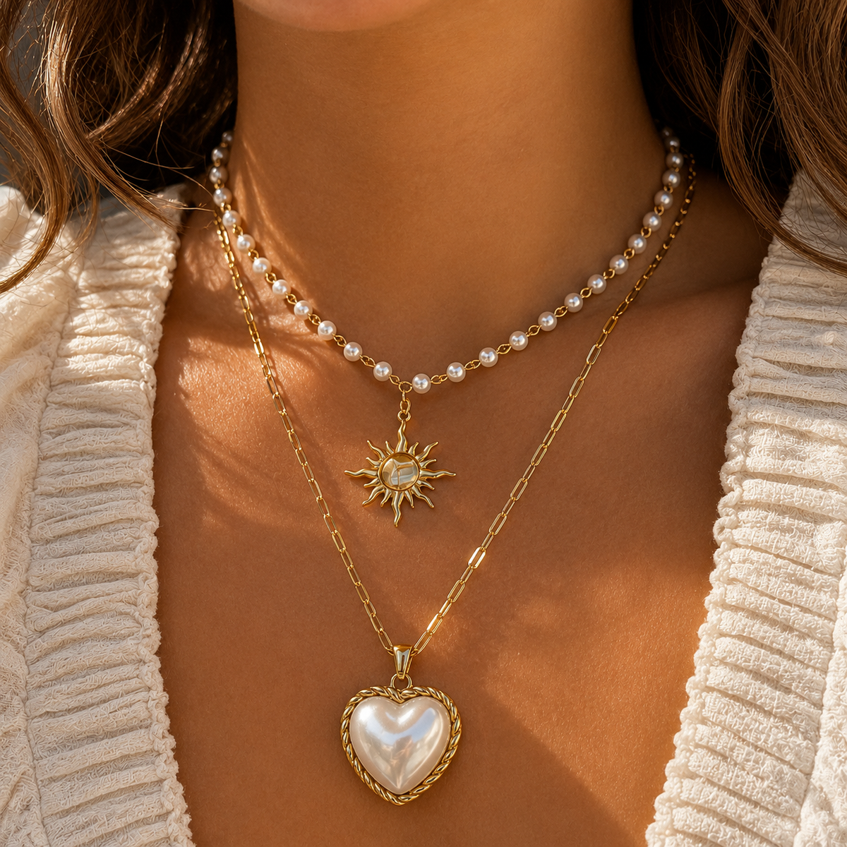 Frau trägt die Perlenkette mit Sonnenanhänger übereinander mit einer langen goldenen Kette mit Perlmutt-Herzanhänger, dazu eine cremefarbene Strickjacke. 3