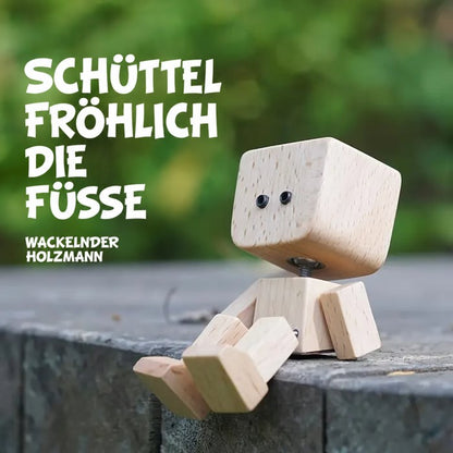 Holzfigur mit schlichten schwarzen Augen sitzt auf einer Steinkante vor grünem Hintergrund und lässt die Füße baumeln. 15