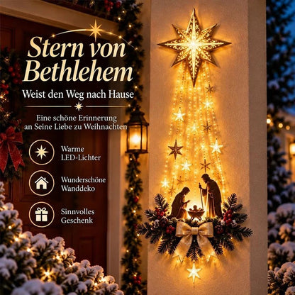 Leuchtender Stern mit Lichterketten und Krippenszene an einer Hauswand neben einer Laterne, umgeben von verschneiten Zweigen 1