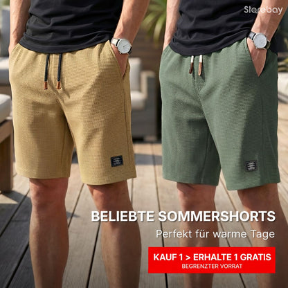 Zwei Männer nebeneinander tragen die Waffelstruktur-Shorts in Sand und Grün zu schwarzen T-Shirts, Hände in den Taschen, auf einer Holzterrasse. 1
