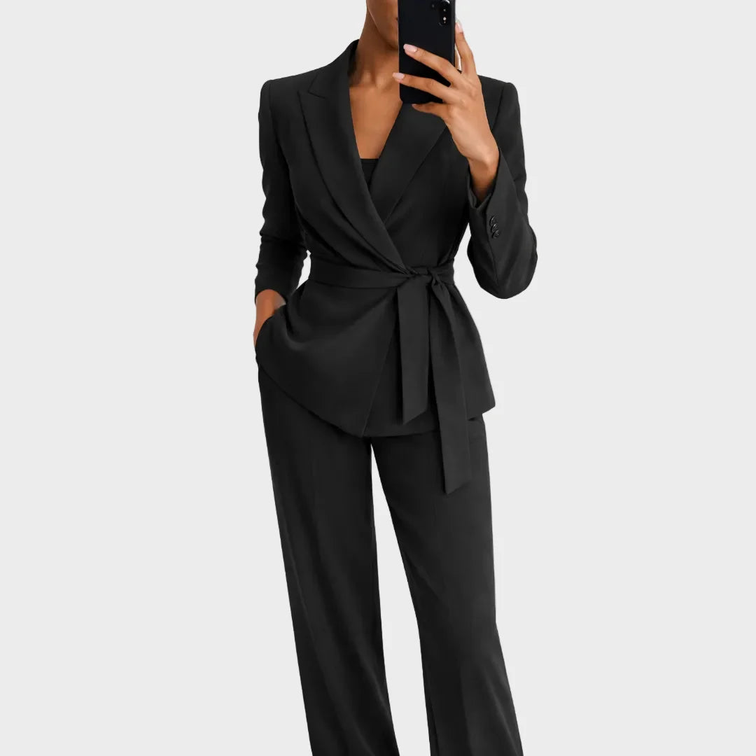 Das Set in Schwarz, Frontansicht: Wickelblazer mit Revers und Bindegürtel zu weiter Hose, die Hand in der Hosentasche.