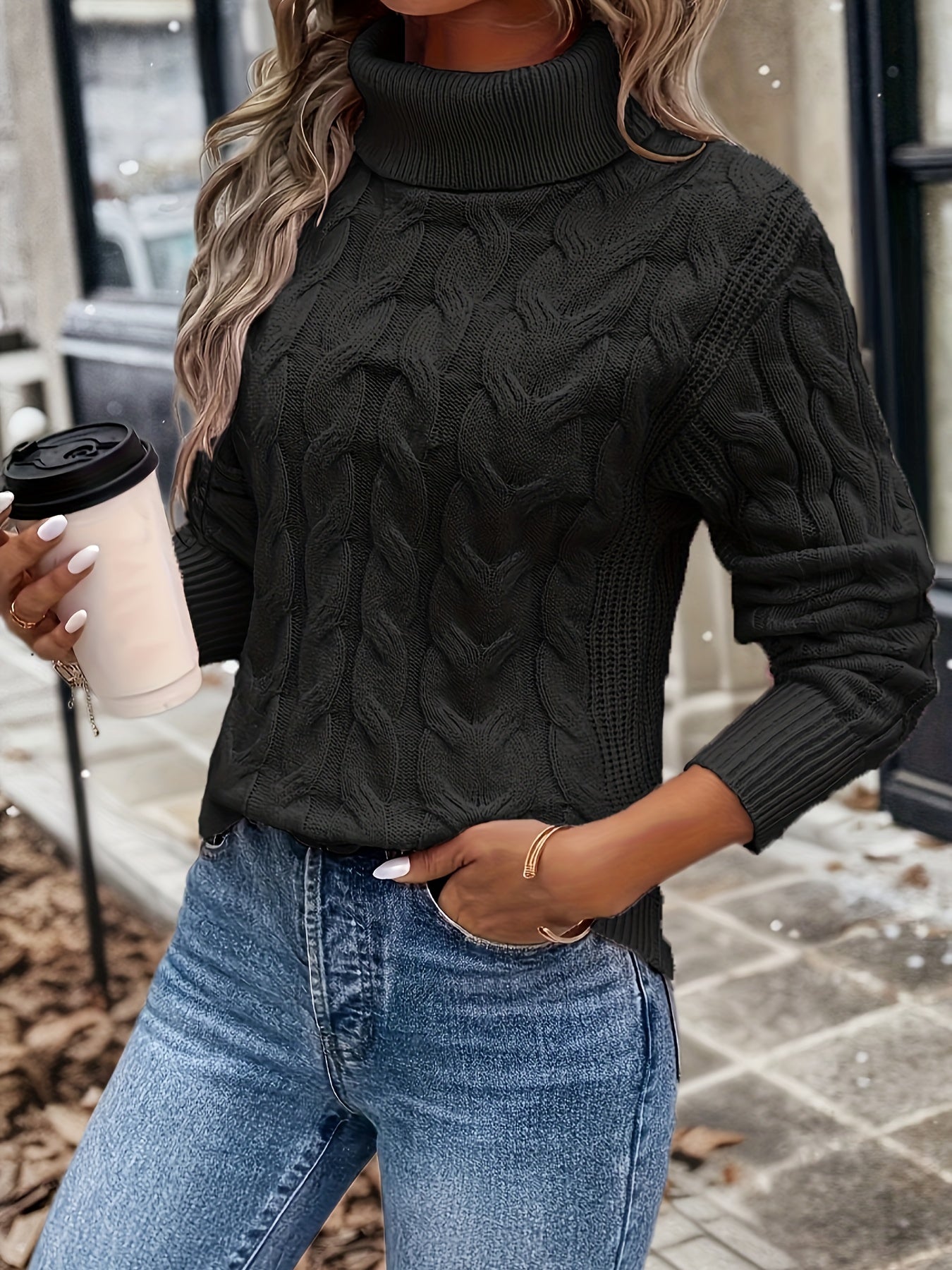 Frau in schwarzem Strickpullover mit Rollkragen, hält Kaffeebecher, trägt blaue Jeans.