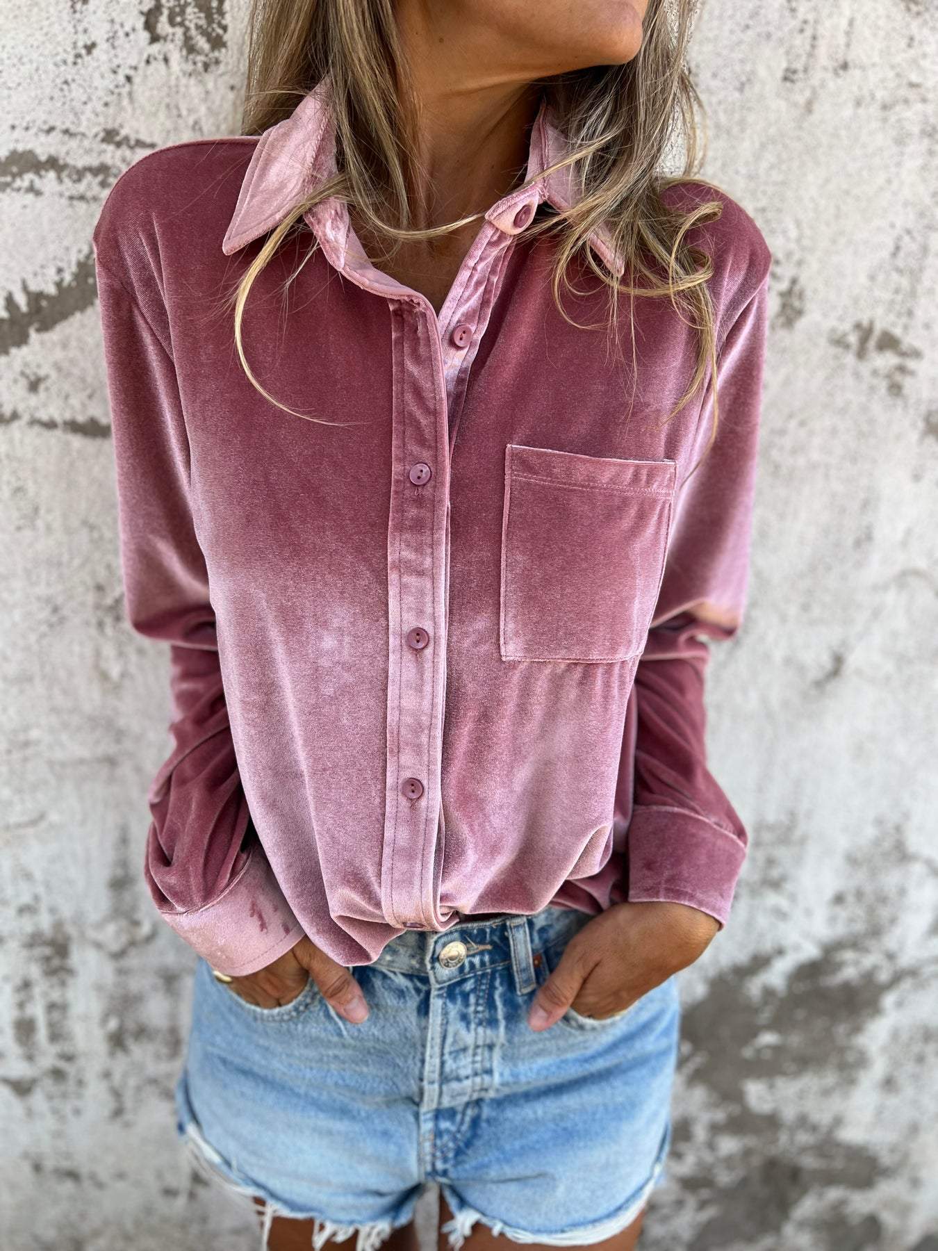 Frau in rosa Samthemd und Jeansshorts vor Betonwand.