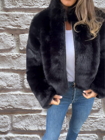 Frau in schwarzer Kunstpelzjacke, weiße Bluse, blaue Jeans vor Ziegelwand.
