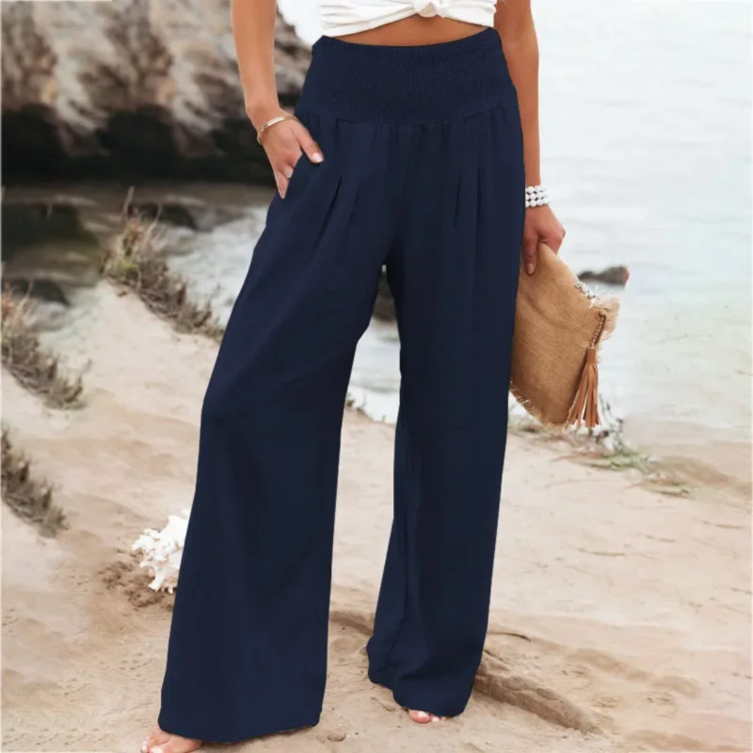 Frau in blauer, weiter Hose am Strand, hält eine beige Tasche, Sommermode, Damenbekleidung.