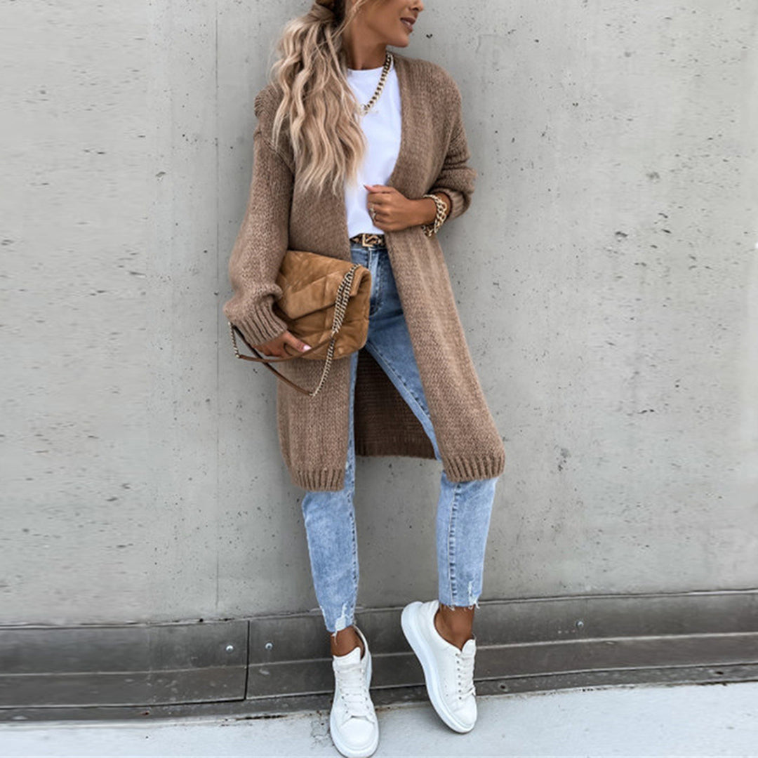 Frau in khakifarbenem Strickmantel, blauer Jeans, weißen Sneakers, brauner Tasche vor Betonwand.