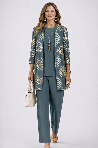 Frau im graublauen Set mit goldenem Palmblattmuster auf dem offenen Cardigan, dazu Rundhalsshirt, weite Hose und cremefarbene Handtasche. 1
