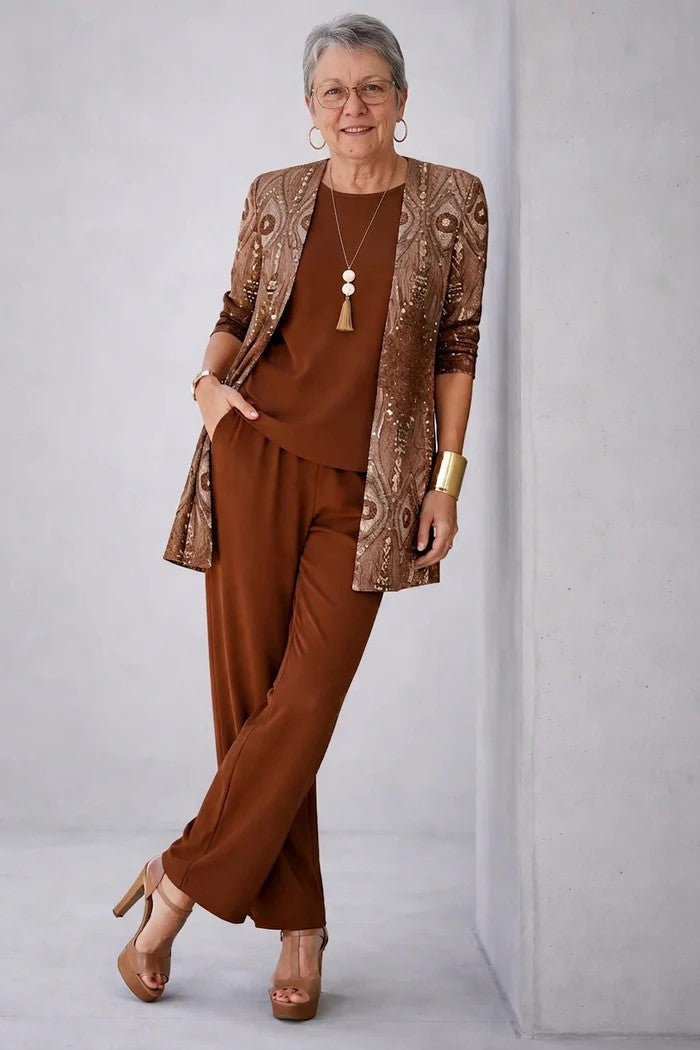 Ältere Frau im braunen Set an eine Betonwand gelehnt, Cardigan mit schimmerndem Ornamentmuster, dazu Shirt und weite Hose. 16
