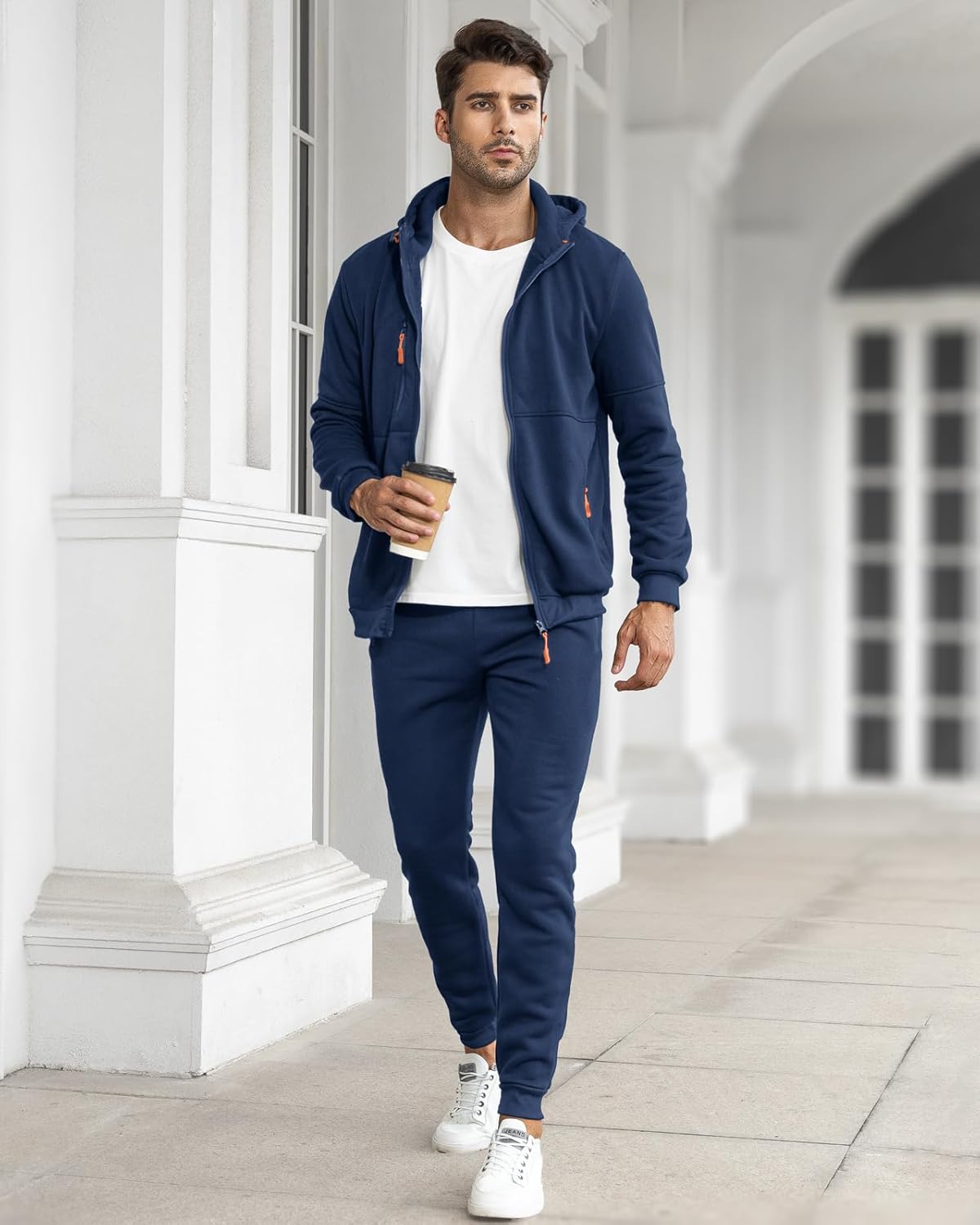 Mann in dunkelblauem Jogginganzug mit Kapuze, hält Kaffeebecher, weiße Sneaker, urbaner Hintergrund.