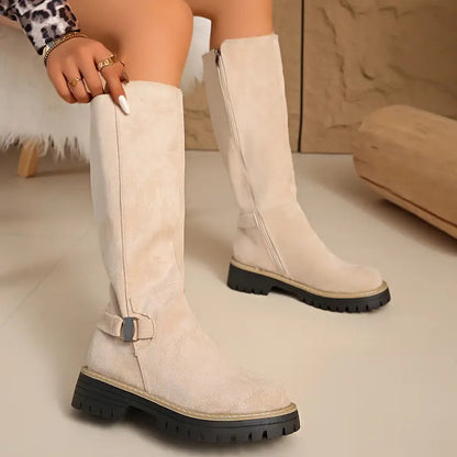 Beigefarbene kniehohe Stiefel im Sitzen, mit Hand am Schaft sowie Schnalle und seitlichem Reißverschluss.